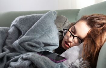Young woman exhausted from smartphone addiction, sleeps on the sofa with her phone. Concept of excessive social media browsing, internet overuse and constant online connection